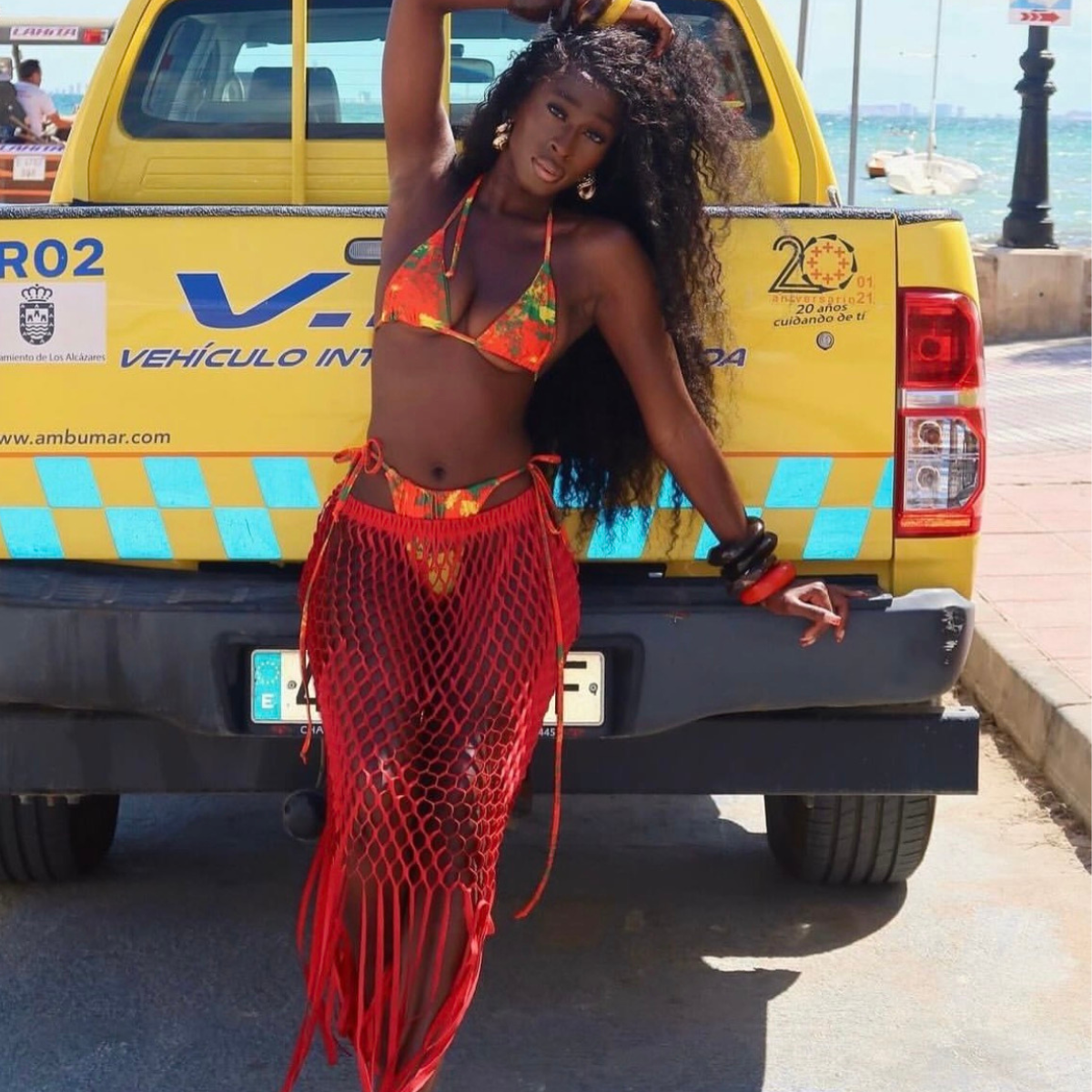 Woman in a colorful bikini and red net skirt standing in front of a yellow vehicle by the beach.