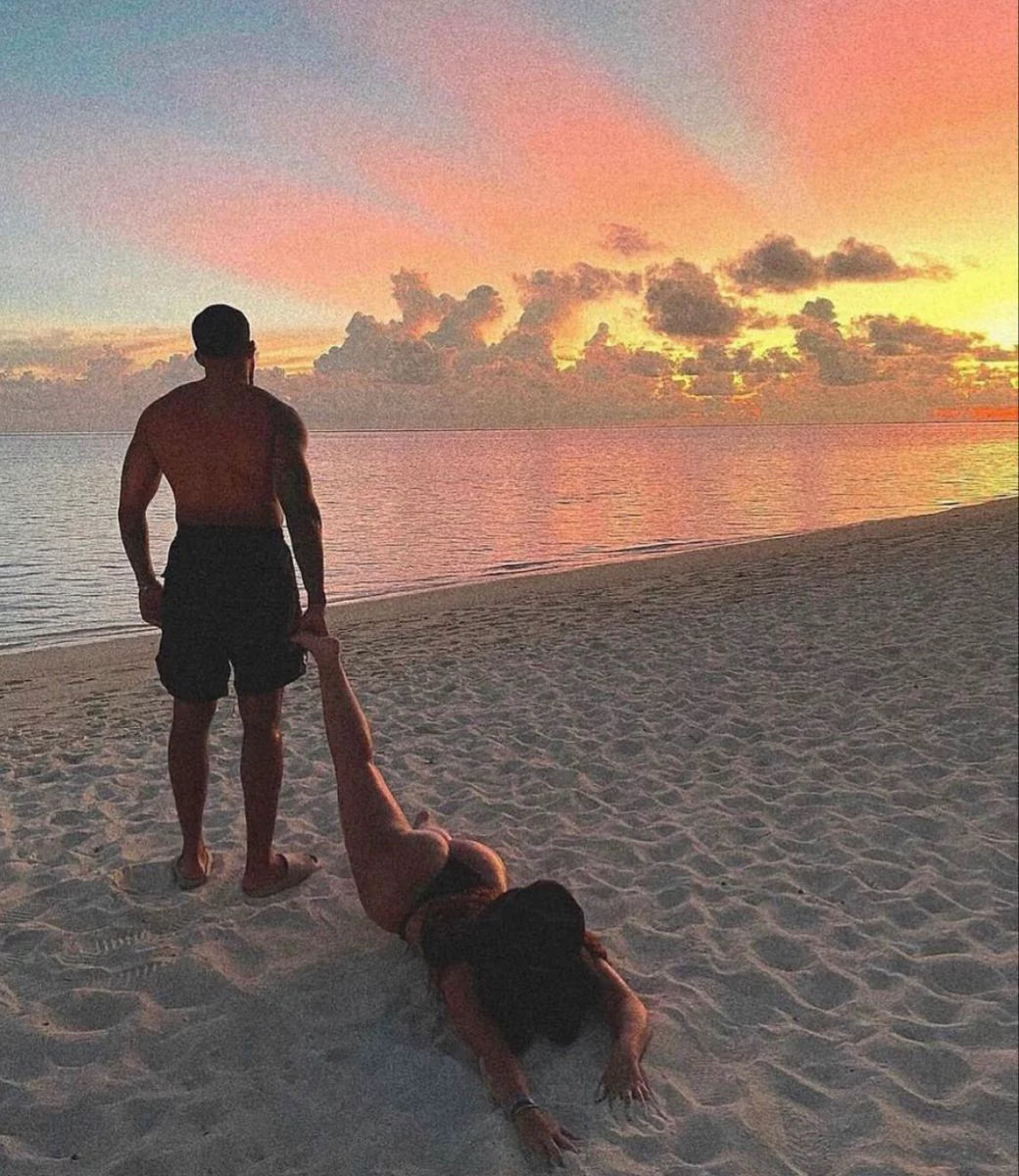 A couple on a beach at sunset, with a man standing and a woman lying on the sand stretching toward the sky. The scene is bathed in warm pink, orange, and yellow sunset light.Belle Exotics is a discreet adult toy and sexual wellness store in Trinidad and Tobago, offering pleasure products, lubricants, lingerie, couple toys, self-love essentials, and intimacy accessories for private online and in-store shopping.   Belle Exotics offers discreet adult toys, lubricants, lingerie, and sexual wellness products in 