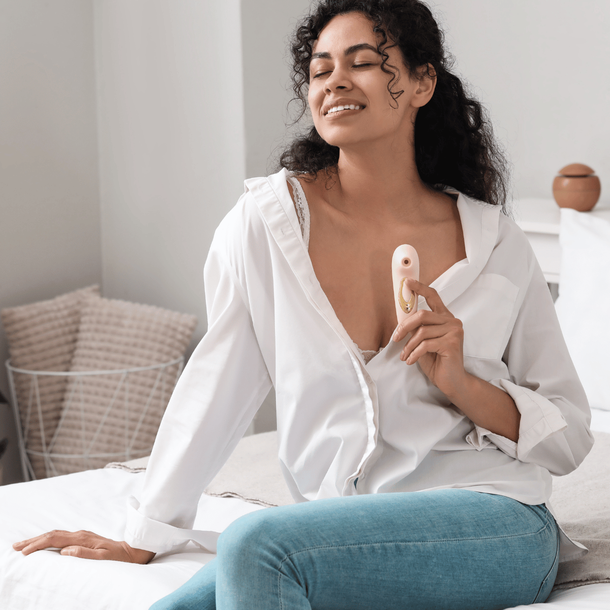 Woman sitting on a bed holding a small female vibrator wearing a white shirt and blue jeans.Pleasuremas 2025 collection featuring luxury adult toys, candles, and intimate gift sets from Belle Exotics.