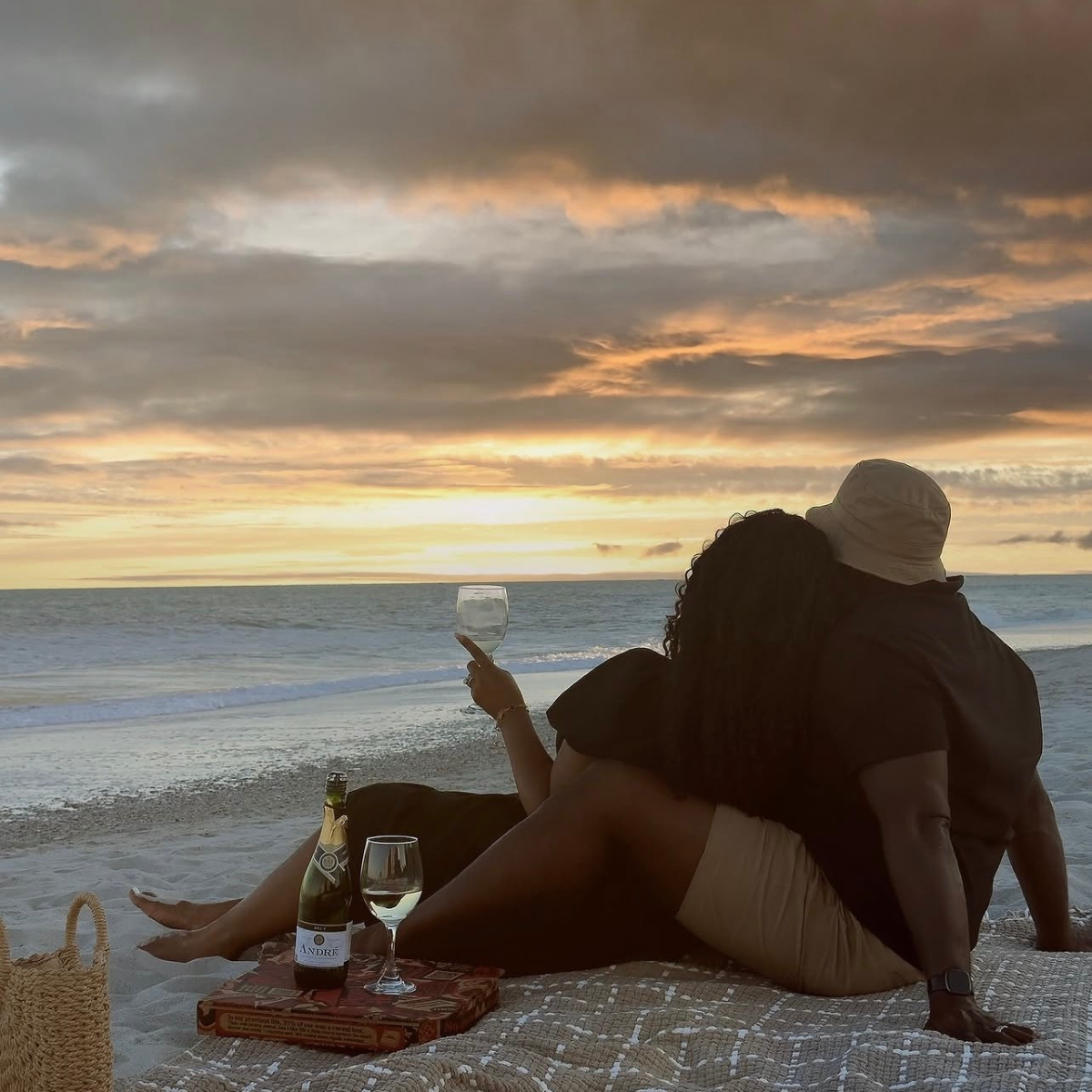 Two people sitting on a blanket at sunset on a beach, enjoying drinks.  Belle Exotics offers discreet adult toys, lubricants, lingerie, and sexual wellness products in Trinidad and Tobago for online and in-store shopping.