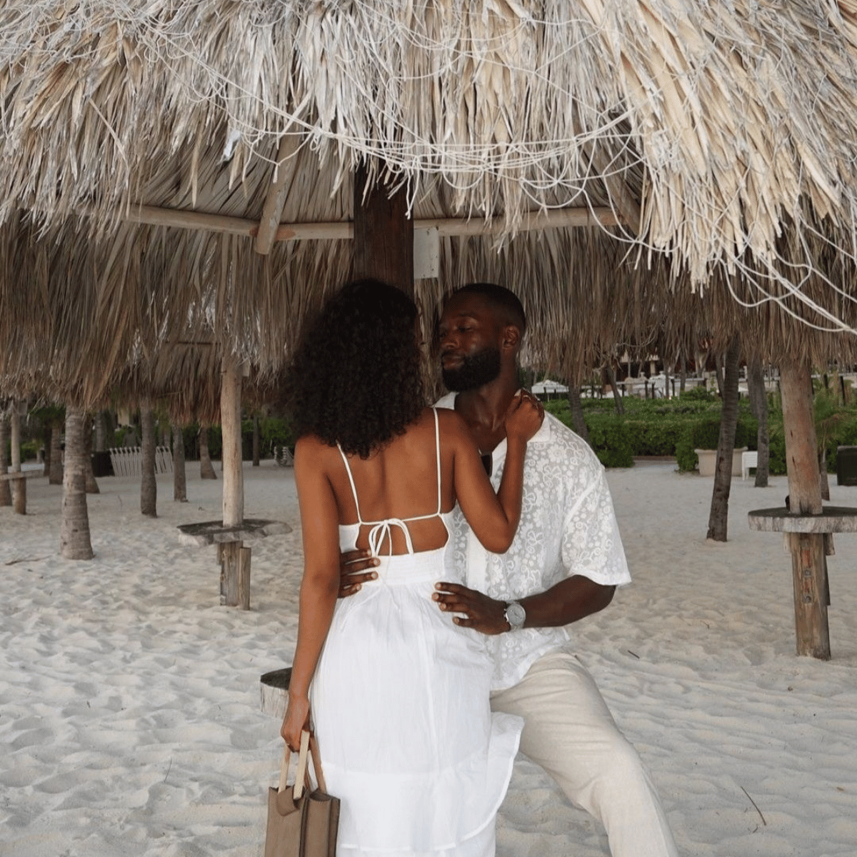 Couple in white outfits sitting under a thatched roof on a sandy beach.