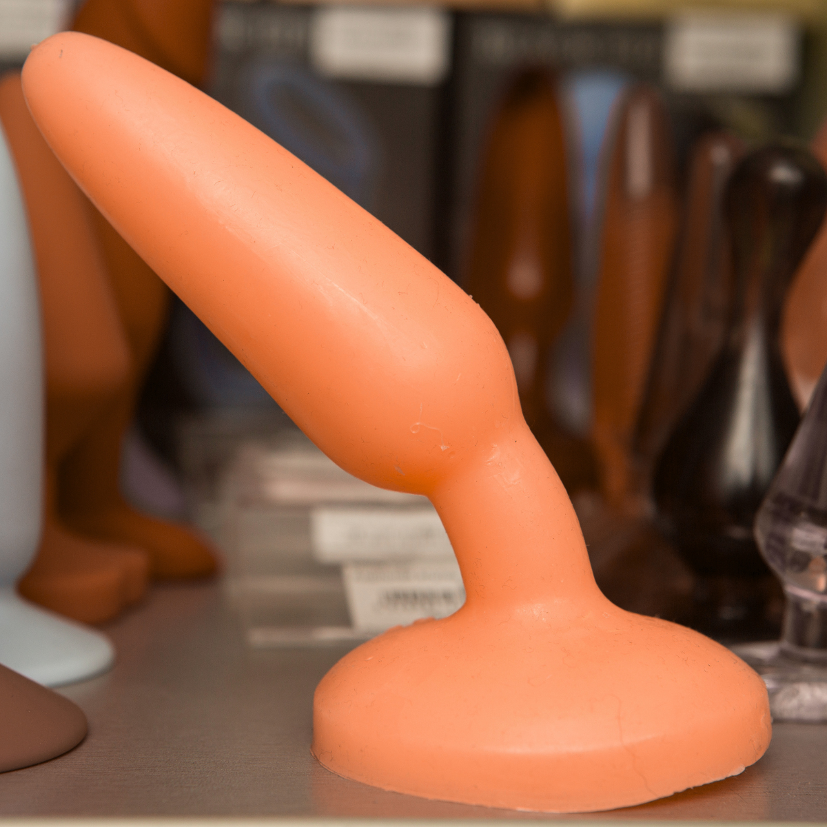 Close-up of an orange silicone anal plug on a shelf with blurred background    Discover pleasure products in Trinidad and Tobago from Belle Exotics, including couple toys, lubricants, lingerie, and beginner-friendly adult product.
