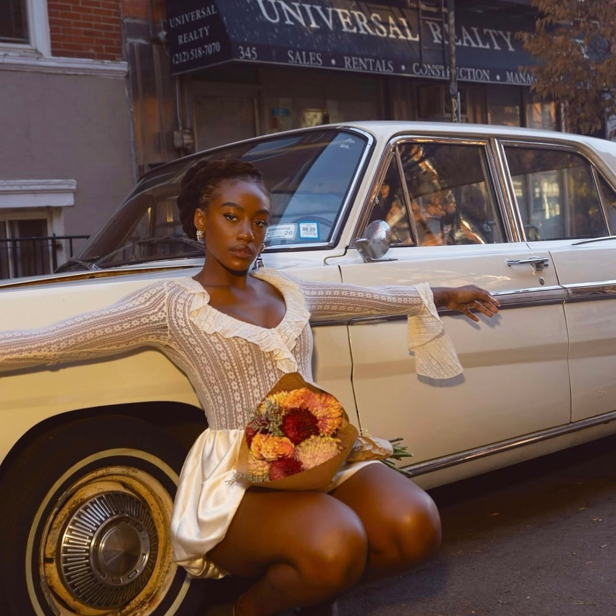 Woman sitting on a vintage car holding flowers, with a building and sign in the background.    Discover pleasure products in Trinidad and Tobago from Belle Exotics, including couple toys, lubricants, lingerie, and beginner-friendly adult products.