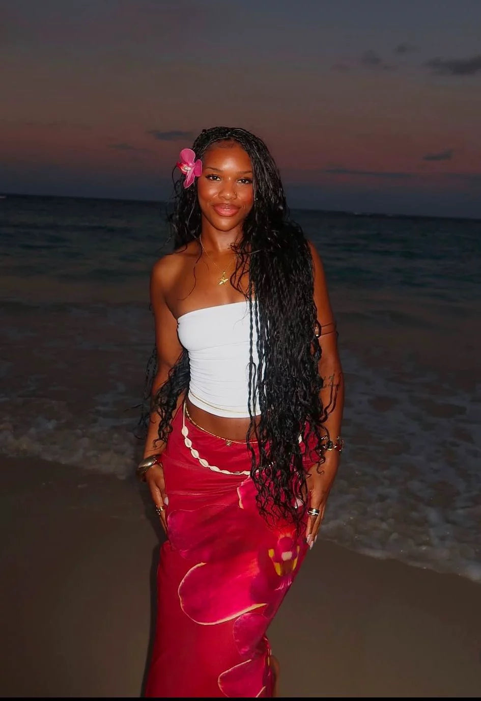 A woman standing on a beach at dusk, wearing a white strapless top and a bright pink wrap skirt with gold trim, with long black braids and a pink flower in her hair.Belle Exotics is a discreet adult toy and sexual wellness store in Trinidad and Tobago, offering pleasure products, lubricants, lingerie, couple toys, self-love essentials, and intimacy accessories for private online and in-store shopping.   
