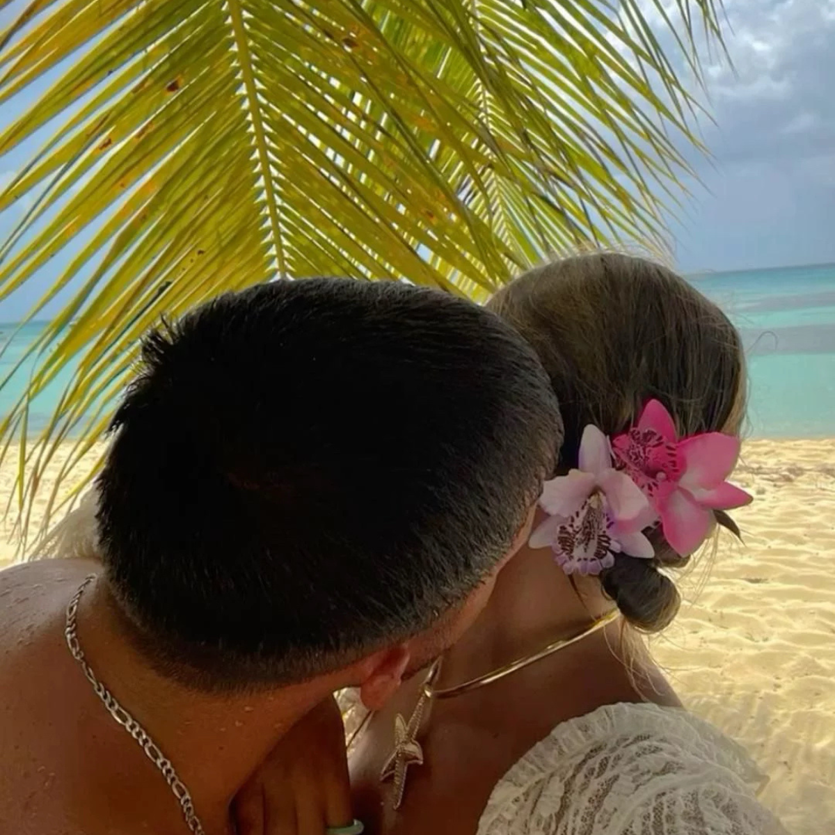 A close-up beach scene showing the back of a man's head and a woman's head in profile, with ocean and palm leaves in the background. The woman has a black bun adorned with pink orchids, and the man wears a silver chain necklace.Belle Exotics is a discreet adult toy and sexual wellness store in Trinidad and Tobago, offering pleasure products, lubricants, lingerie, couple toys, self-love essentials, and intimacy accessories for private online and in-store shopping.   Belle Exotics offers discreet adult toys, 