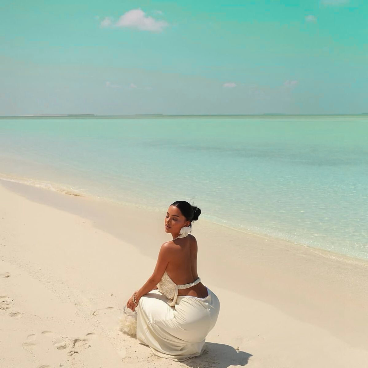 Woman in a white dress sitting on a sandy beach with clear turquoise water and sky.Your Golden Hour campaign by Belle Exotics in Trinidad and Tobago promoting confident exploration, sexual wellness, and guided intimacy in-store and online.