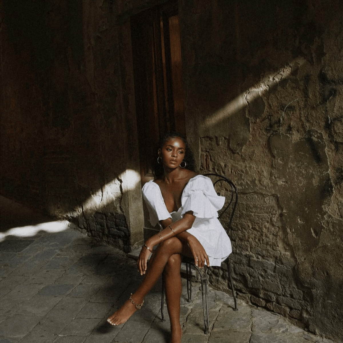 A woman sits on a black metal chair in a rustic stone corridor, wearing a white ruffled short dress and gold jewelry.Shop Belle Exotics for intimacy, pleasure and adult wellness products in Trinidad & Tobago. Shop online or visit our Marabella and Valsayn locations.