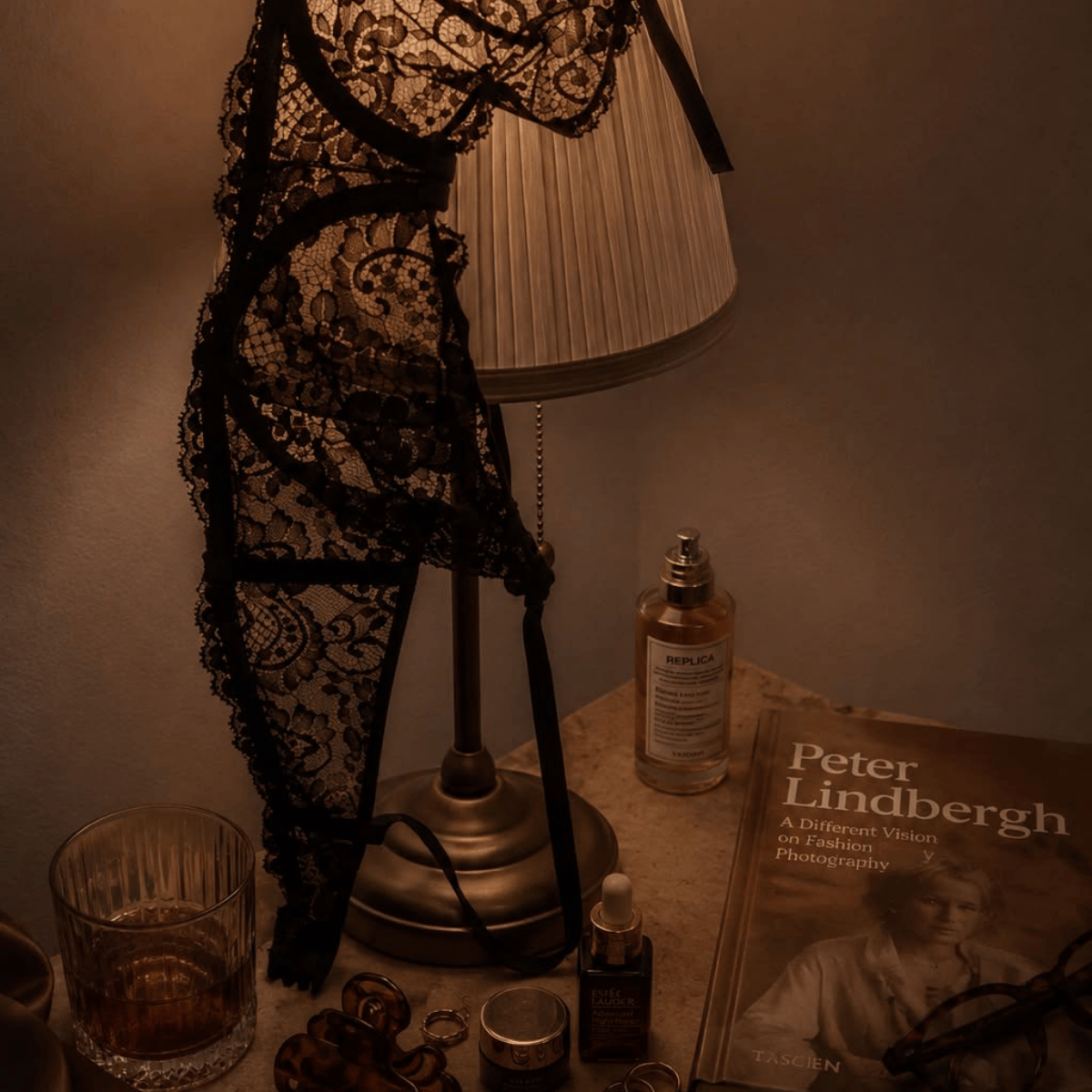 A black lace lingerie bodysuit draped over a table lamp on a dimly lit tabletop, surrounded by a glass, a perfume bottle, a small jar, and a Peter Lindbergh book.Shop Belle Exotics for intimacy, pleasure and adult wellness products in Trinidad & Tobago. Shop online or visit our Marabella and Valsayn locations.