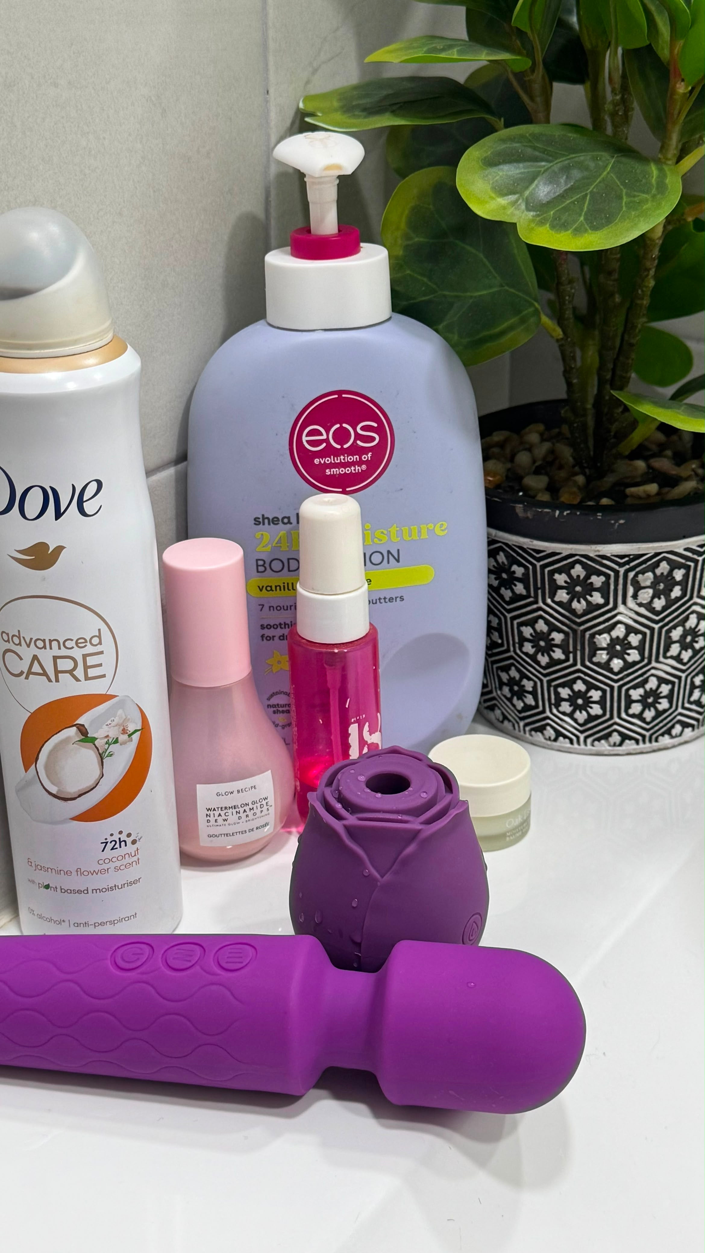 A purple handheld personal massager with a flower-shaped head attachment, displayed alongside various personal care products including a Dove body wash bottle, an EOS body lotion bottle, and a pink spray bottle on a white surface next to a potted plant.Belle Exotics is a discreet adult toy and sexual wellness store in Trinidad and Tobago, offering pleasure products, lubricants, lingerie, couple toys, self-love essentials, and intimacy accessories for private online and in-store shopping. 